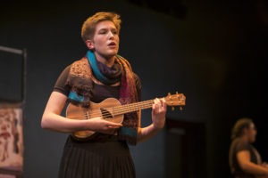 Young white woman with short cropped brown hair strums a ukulele and sings.