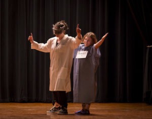Two young white children on stage. The one on the left is wearing a white lab coat and has her hands raised with her index finger pointing up. The shorter one behind is wearing a light blue hospital johnnie and has ber hands raised, palms facing up.