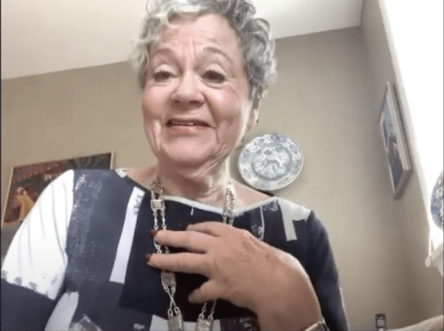 Older white woman with short cropped white haor smiles down at the camera. Her hand is on her chest. She is wearing a geometric print blouse in navy blue and white.