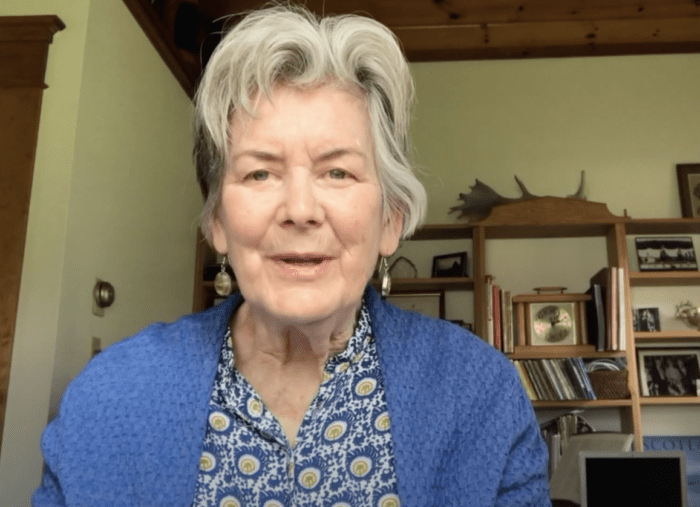 An older white woman with short cut white hair speaks directly to the camera. She is wearing a blue cardigan over a blue print blouse and dangling silver earrings.
