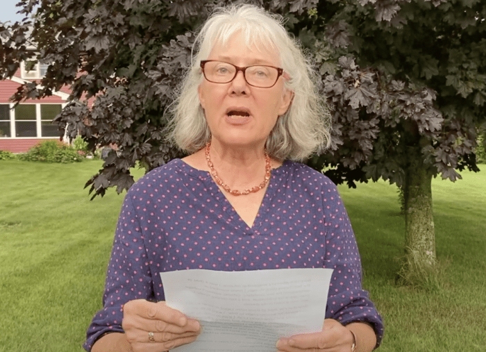 Older white woman with shoulder length white hair and eyeglasses stands in front of a tree in full leaf, holding a piece of paper and speaking to the camera.