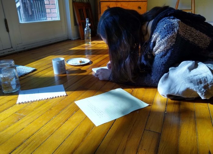 A young person with long dark hair that covers their face crouches on a hardwood floor writes by hand. They are surrounded by papers and a mason jar, coffee mug, clear glass bottle, and a plate with one piece of food left on it. STrong sunlight streams in through a glass door at left.