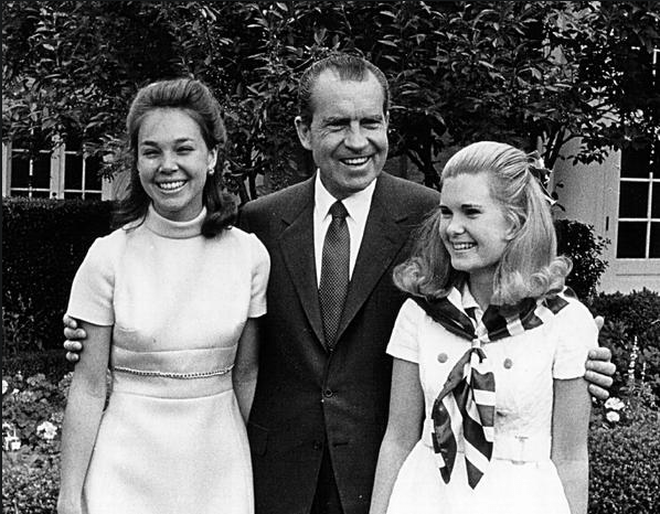 US President Richard M. Nixon with his daughters Julie and Tricia. He is wearing a dark suit and tie. The women are both wearing white, short sleeved dresses. They are all smiling.