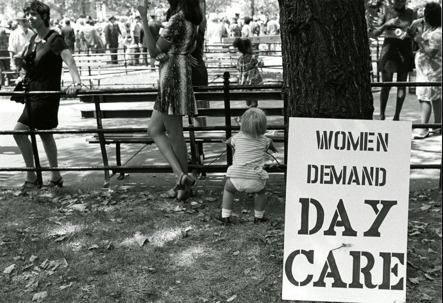 A sign leaning against a tree reads "Women Demand Day Care." A few small children and many women are seen in the background.