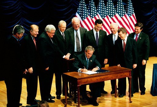 US President George W. Bush wearing a dark suit and tie is seated at a table signing the partial abortion ban bill. Nine middle aged and elderly white men in suits are standing behind him. Six poles with American flags hang in the background.