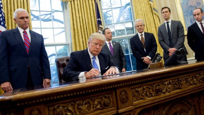 US President Donald J. Trump seated at the Resolute desk in the oval office. Vice President Mike Pence stands to his right, and four other white men stand to his left. All the me are wearing dark suits and ties.