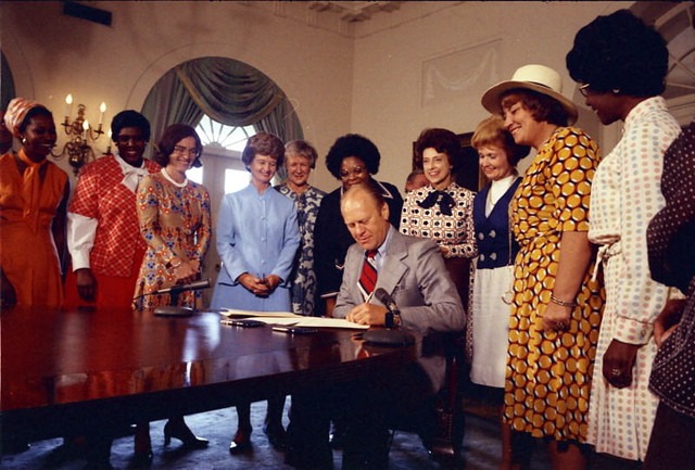 US President Gerald Ford signing a bill into law. He is surrounded by women of many races, including his wife Betty, Bella Abzug, and Shirley Chisholm.