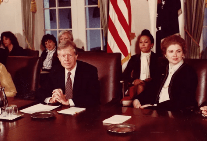 US President Jimmy Carter sits at a desk in a conference room. All the rest of the people in the photo are woman.