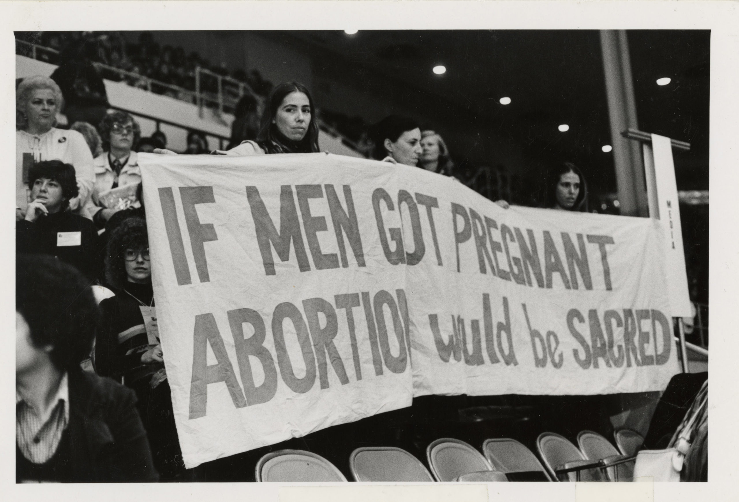 White women in a stadium holding a large banner reading "If men got pregnant abortion would be sacred."