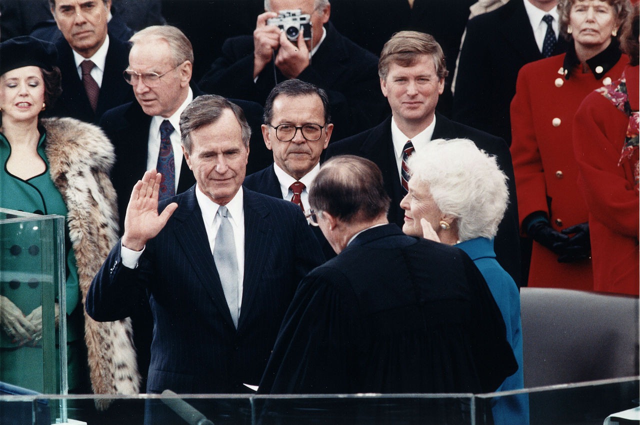US President George H. W. Bush takes the oath of office at his inauguration. His hand is raised. His wife Barbara is by his side, and Vice President Dan Quayle is among many other in the background.