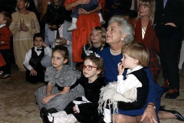 First Lady Barbara Bush wearing a blue dress and her trademark pearl necklace, sitting on the floor with several children. They appear to be watching or listening to a reading or performance.