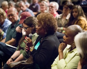 A seated audience of mixed races and ages. In the foreground a Black woman with a light brown Afro is holding a microphone and speaking earnestly.