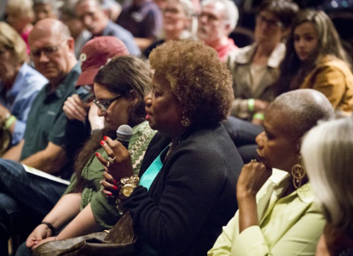 A seated audience of mixed races and ages. In the foreground a Black woman with a light brown Afro is holding a microphone and speaking earnestly.
