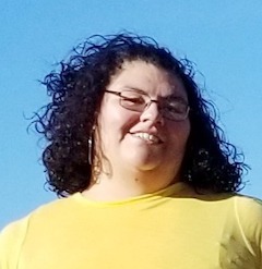 Smiling young indigenous woman with curly shoulder length brown hair. She is wearing eyeglasses and a light yellow top.